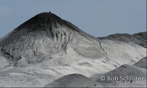 Klein silhouet van een kraai boven op een grote berg grijs zand.