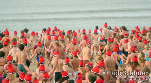 Een groep mensen in badkleding en een oranje muts op rent over het strand naar zee.