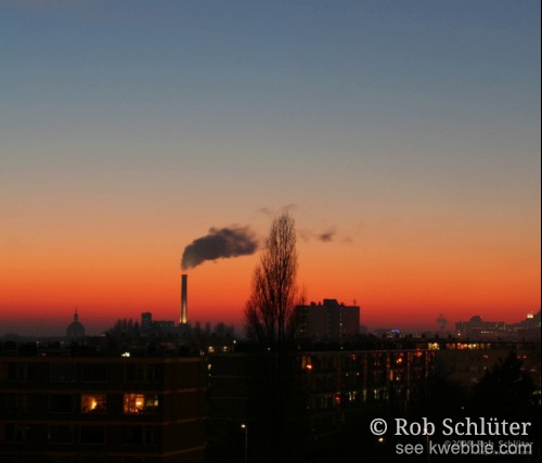 De ondergaande zon kleurt de lucht oranjerood boven de stad Leiden met o.a. de schoorsteen van de energiecentrale.