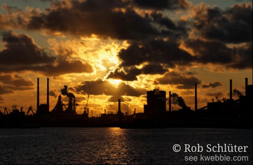 Oranje lucht met de laagstaande zon achter donkere wolken van de lucht en uit silhouetten van schoorstenen en fabrieksgebouwen van Tata Steel. Op de voorgrond het donkerblauwe water van het Noordzeekanaal.