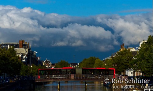 Onder een dreigend beschenen wolkenlucht steekt een gelede rode R-net bus de Nassaubrug over de Nieuwe Gracht over.