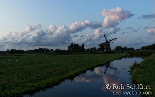 Hollands landschap met wolkenlucht boven een weiland en daartussen een dijk met een windmolen.