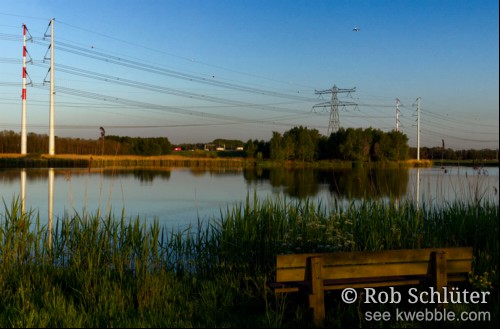 Een bank aan de rietoever van een meertje met uitzicht op hoogspanningsmasten, snelweg en een opstijgend vliegtuig.