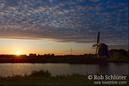 Een windmolen op de oever aan de overkant van een breed kanaal steekt donker af tegen de oranje gloed van de avondzon die nog net over de horizon straalt. De lucht is gevuld met lichte en donkere wolkjes in een regelmatig patroon tegen de donkerblauwe kleur van de avond.